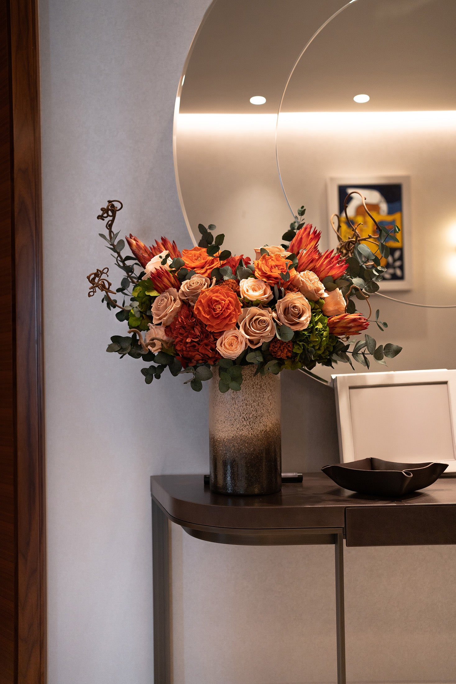 Decorative preserved mixed flowers arrangement in a vase on a table with a mirror reflecting a room interior.