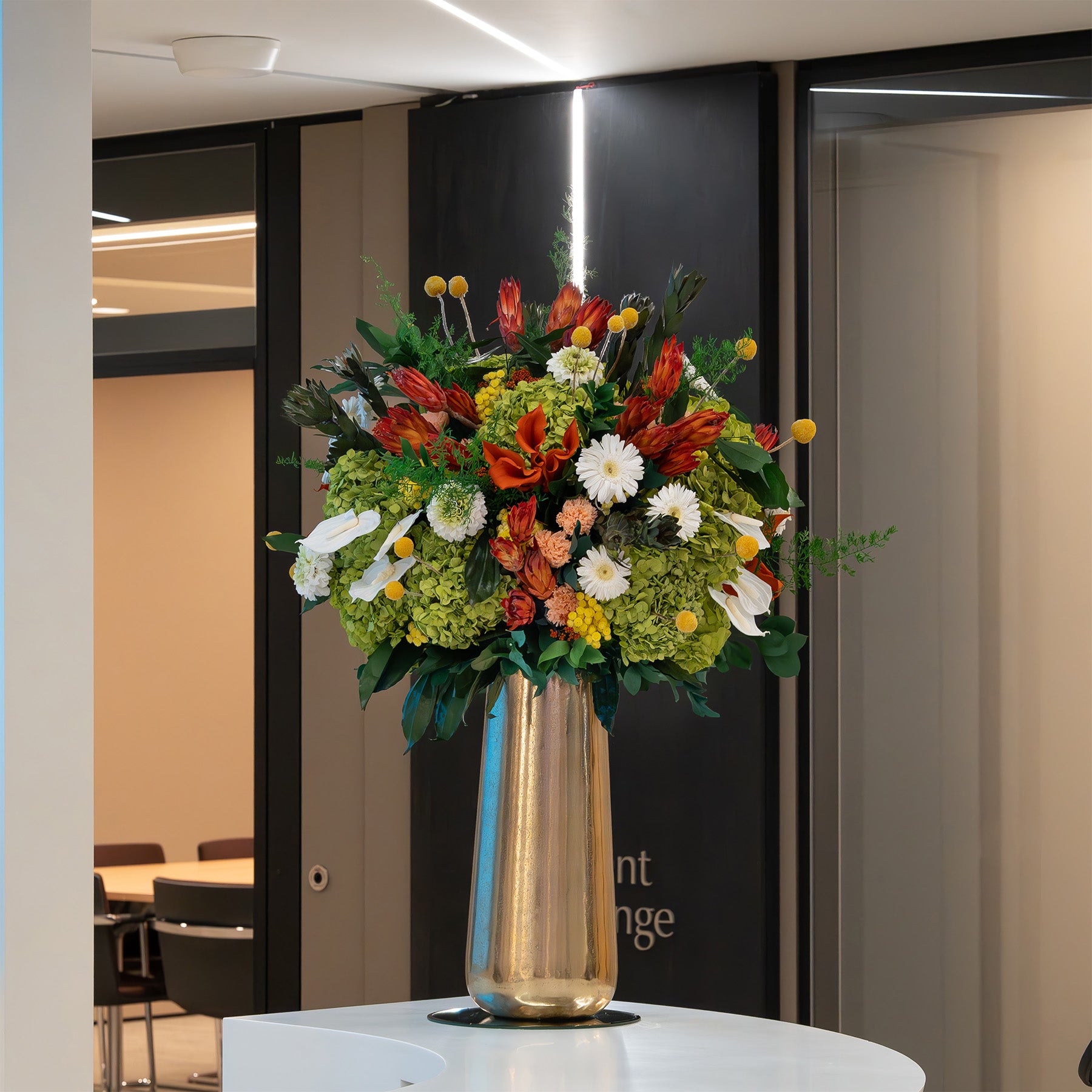 A large, vibrant preserved floral arrangement featuring preserved green hydrangeas, preserved gerberas, anthurium, carnations and dahlia, finished with preserved proteas, leucadendron, in a tall gold vase, displayed on a white pedestal in a modern office lobby.