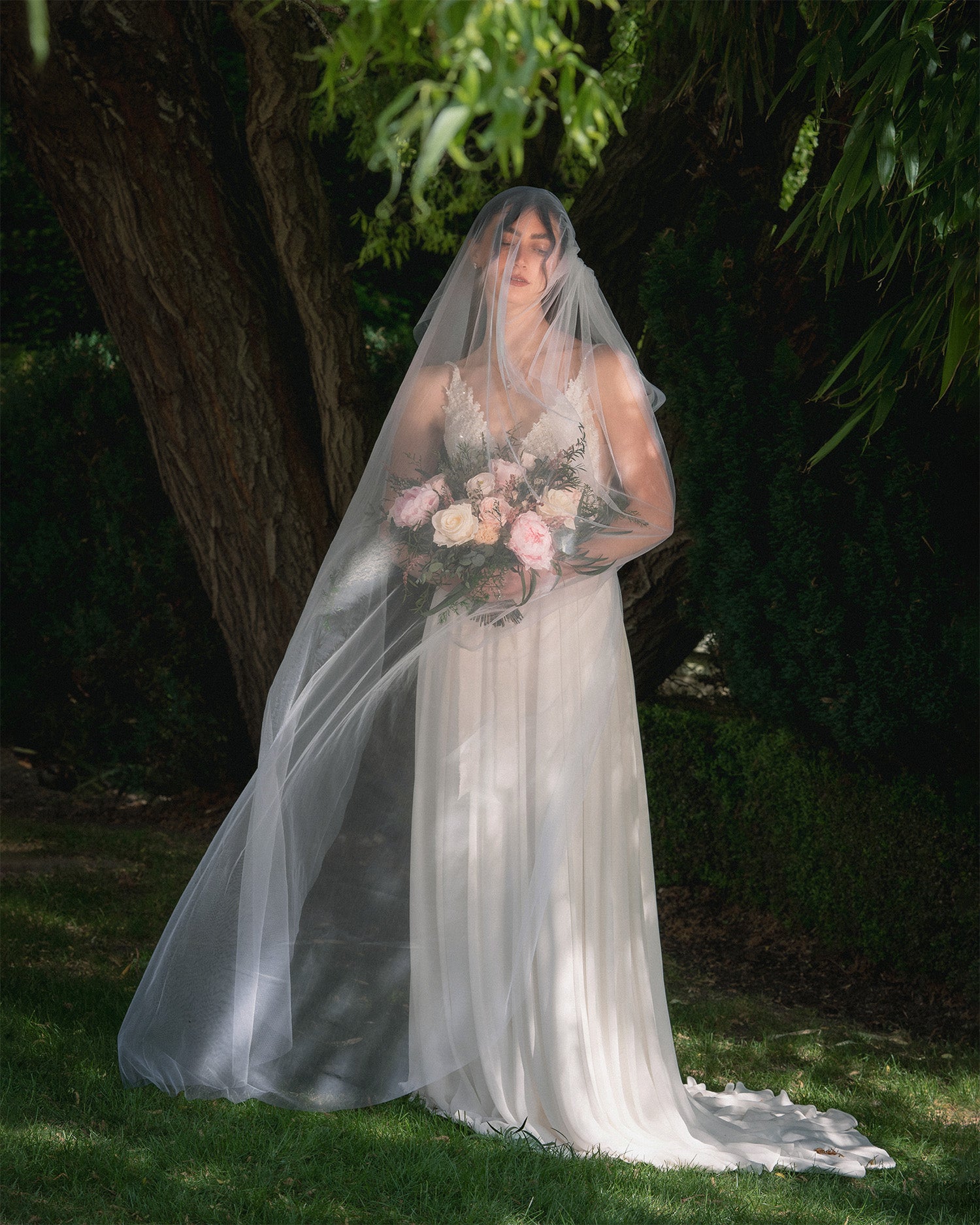 A bride in a wedding gown with a long sheer veil holds a pastel preserved bouquet of pink, peach, and white flowers while standing outdoors in front of a large tree.