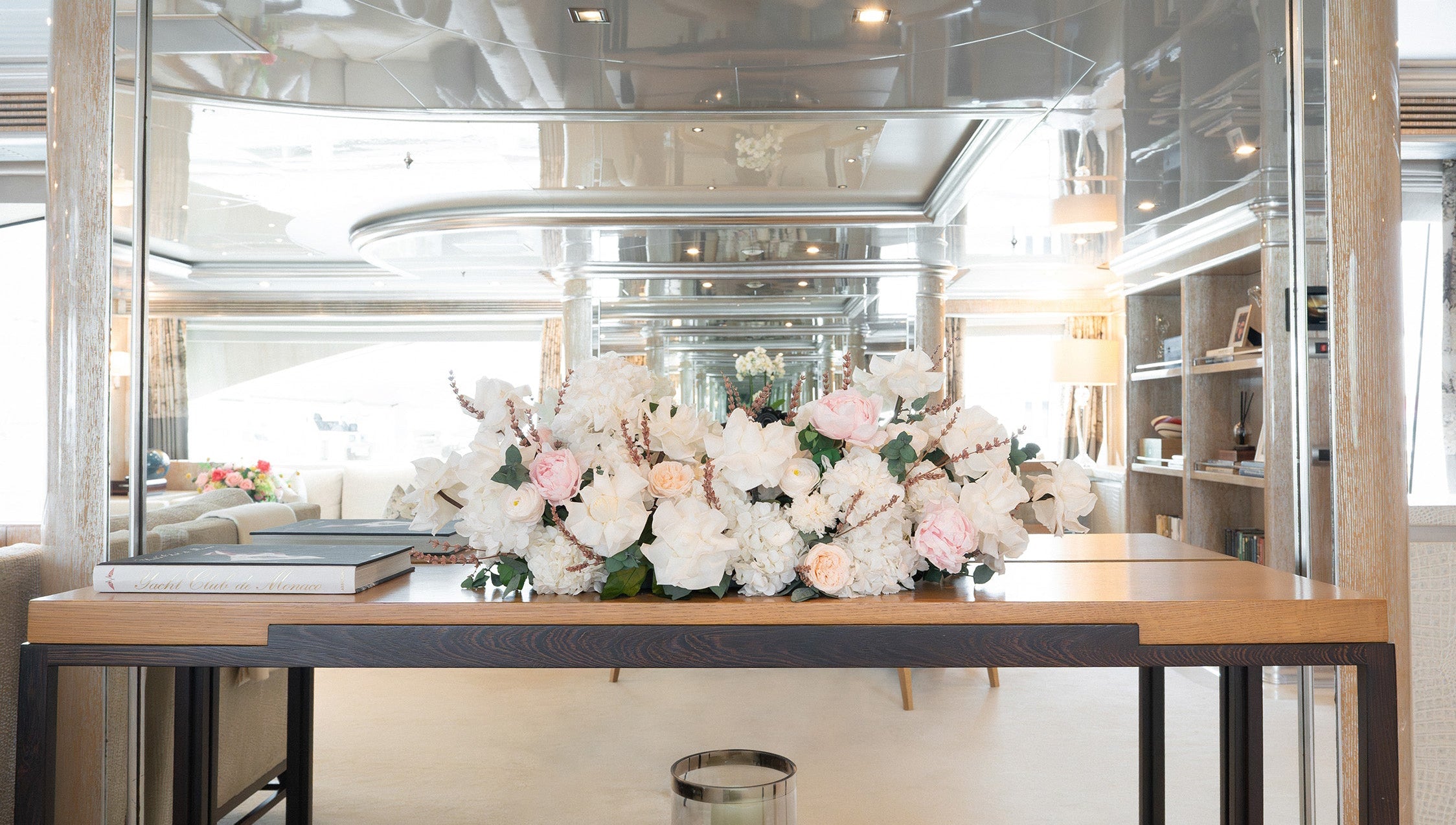 A large preserved floral arrangement of white roses and light pink peonies with green foliage, displayed on a wooden table on board a yacht.