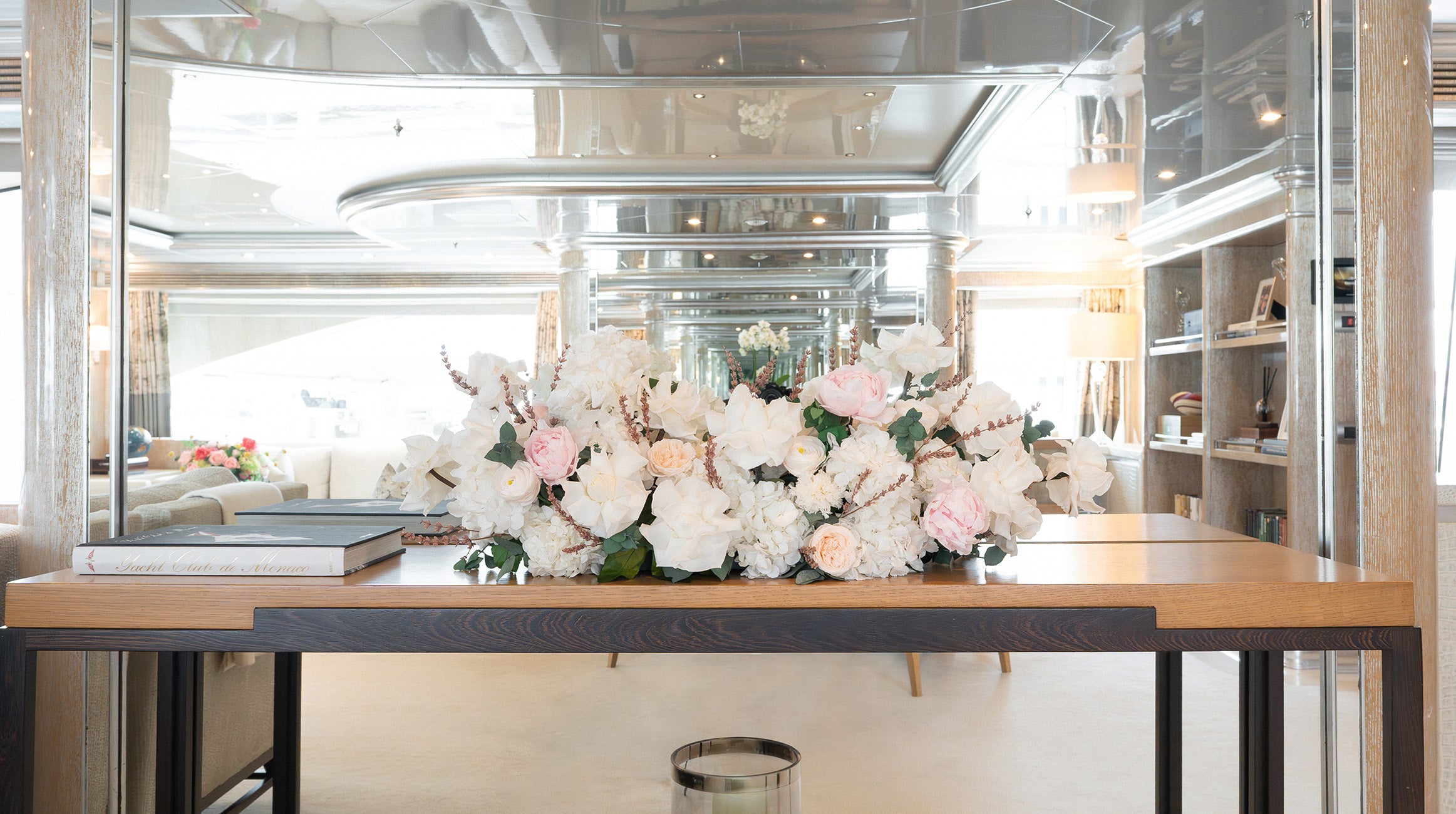 A large preserved floral arrangement of white roses and light pink peonies with green foliage, displayed on a wooden table on board a yacht.