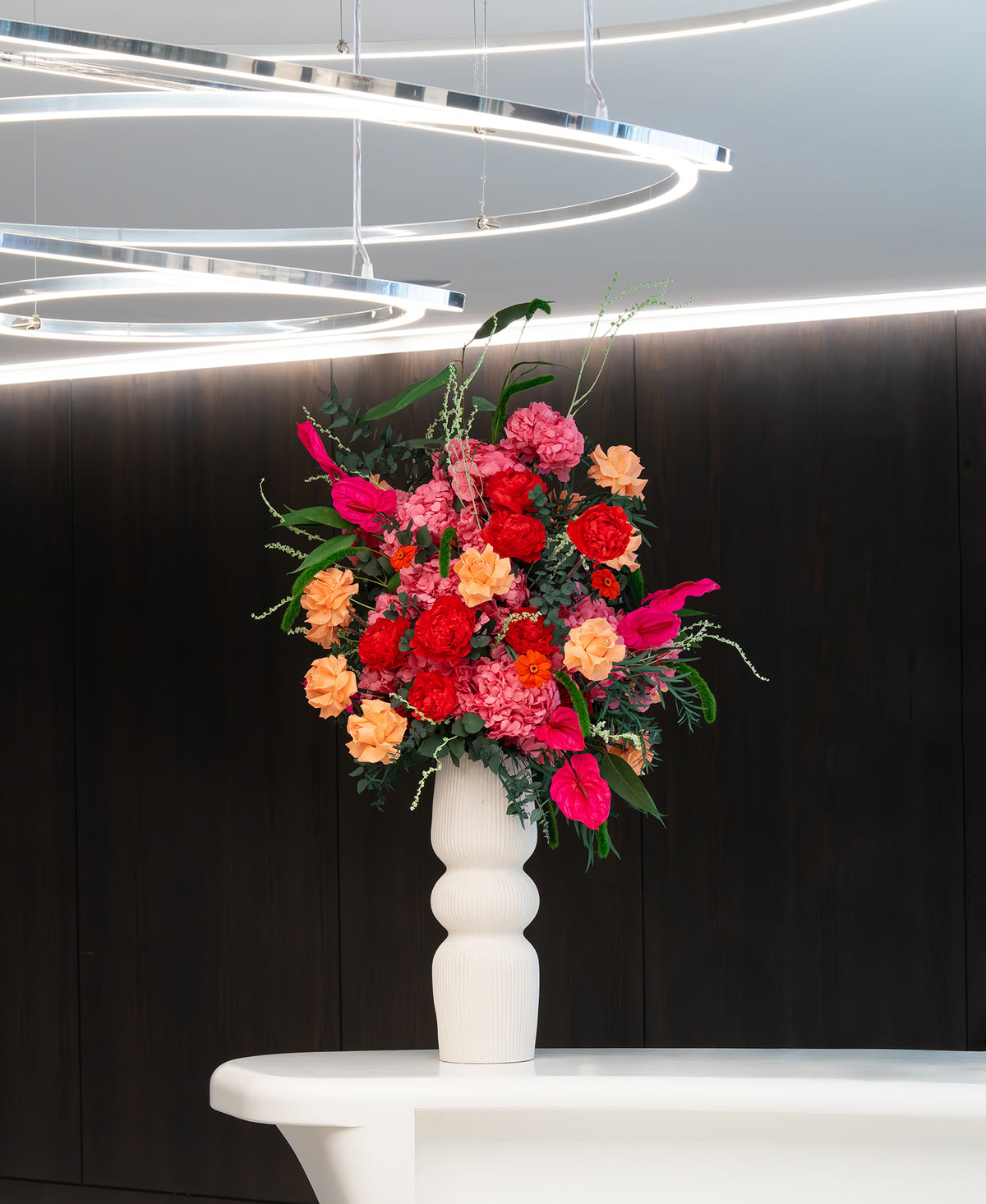 A preserved vibrant floral arrangement featuring preserved red peonies, preserved hot pink anthurium, preserved orange roses, and preserved pink hydrangeas with green foliage, presented in a white sculptural vase on a white table in a office space. 