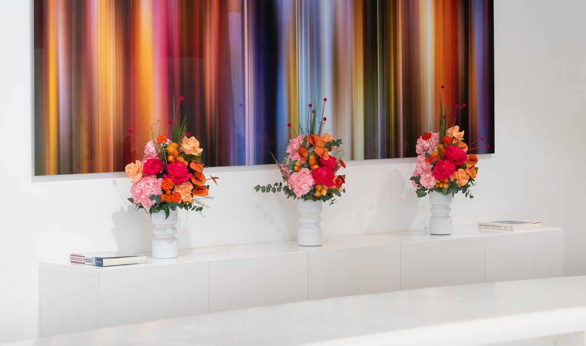 Three white vases with preserved pink hydrangeas, preserved hot pink peonies, orange preserved reflexed flowers and filler flowers. The floral arrangements sits on the console in a bright, modern room.