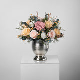 A preserved blush and peach gardenia and roses, gardenia arrangement with foliage in silver vase