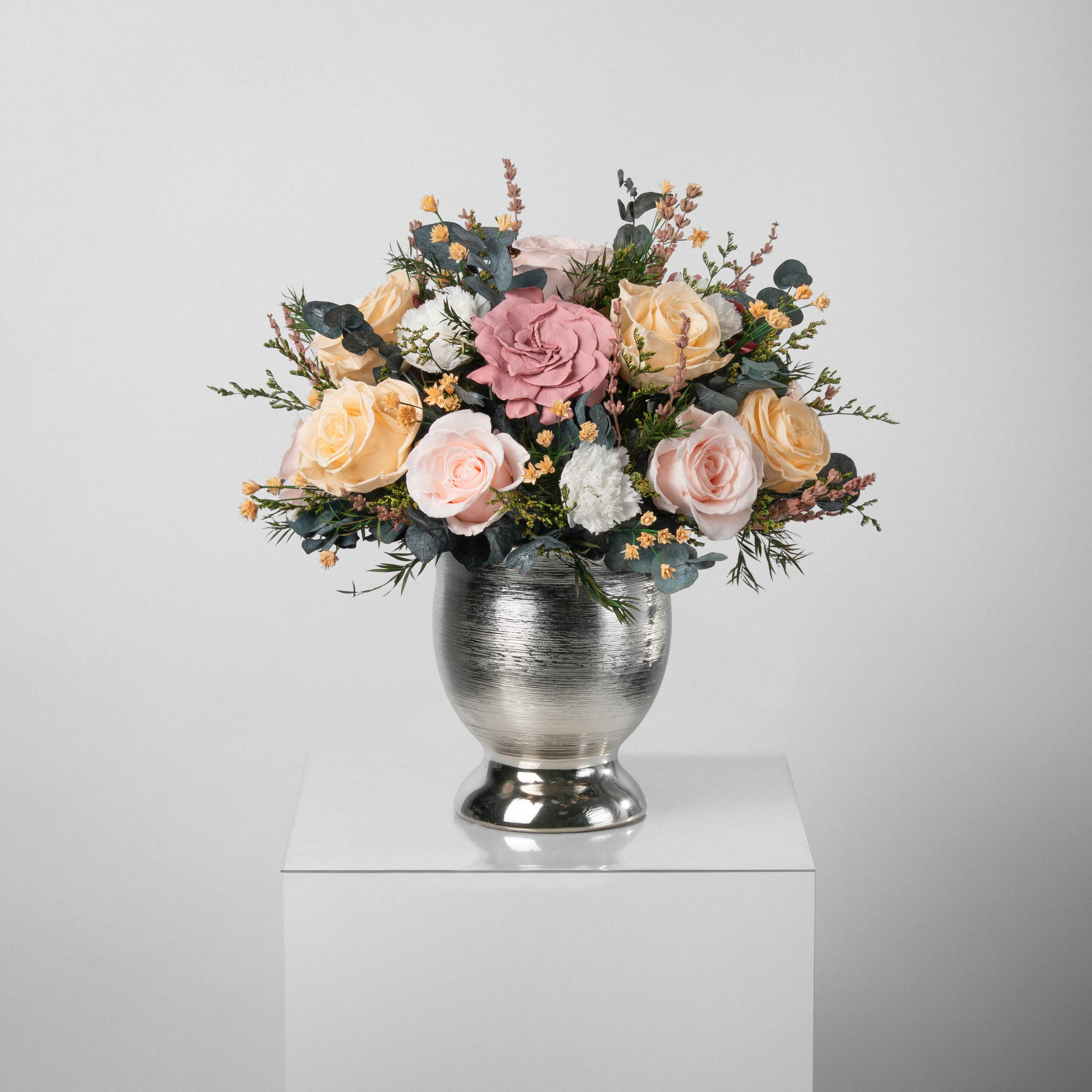 A preserved blush and peach gardenia and roses, gardenia arrangement with foliage in silver vase