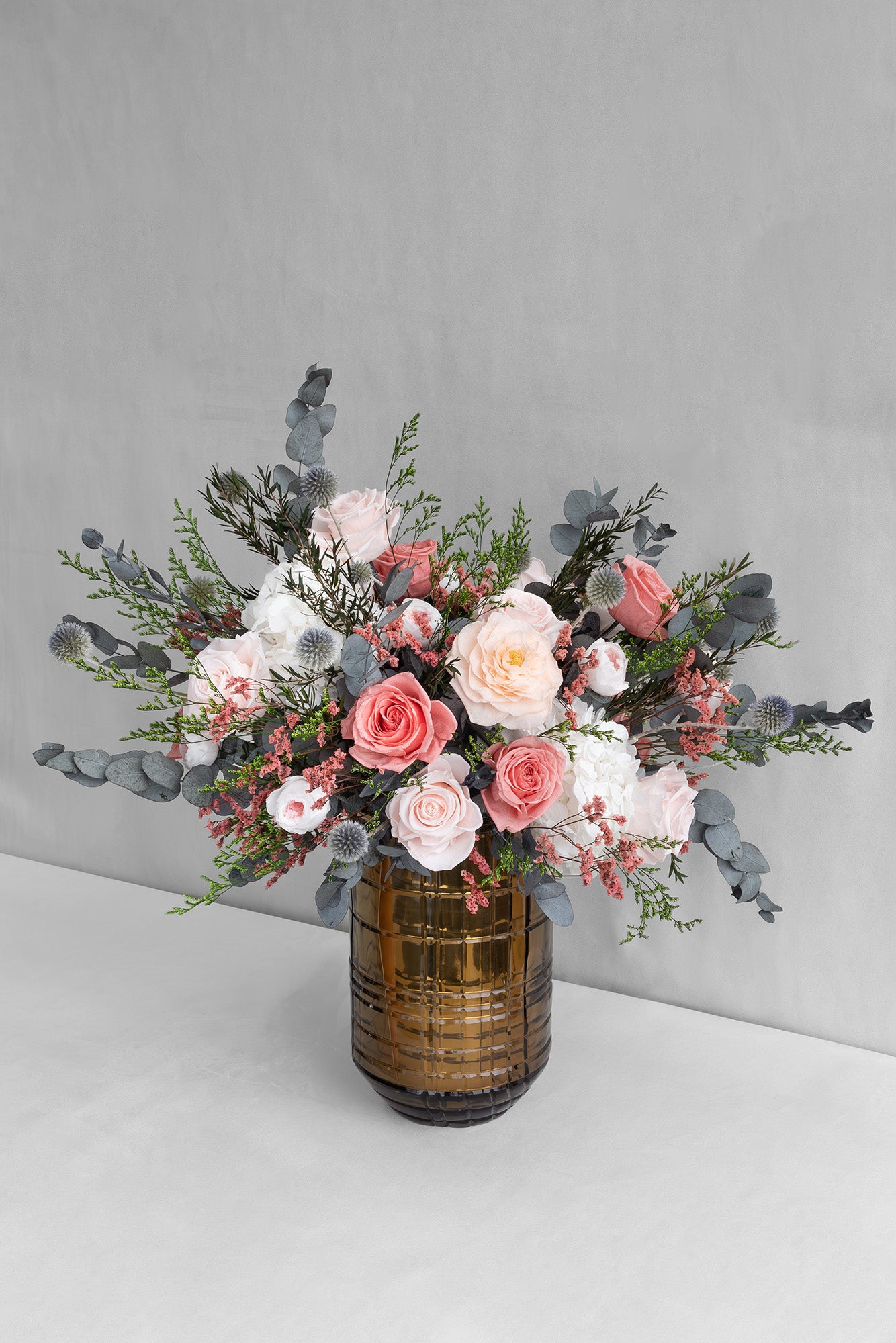 Bouquet of preserved pink and blush roses, white hydrangeas and thistles with tartarica and preserved eucalyptus in a decorative vase on a light grey background