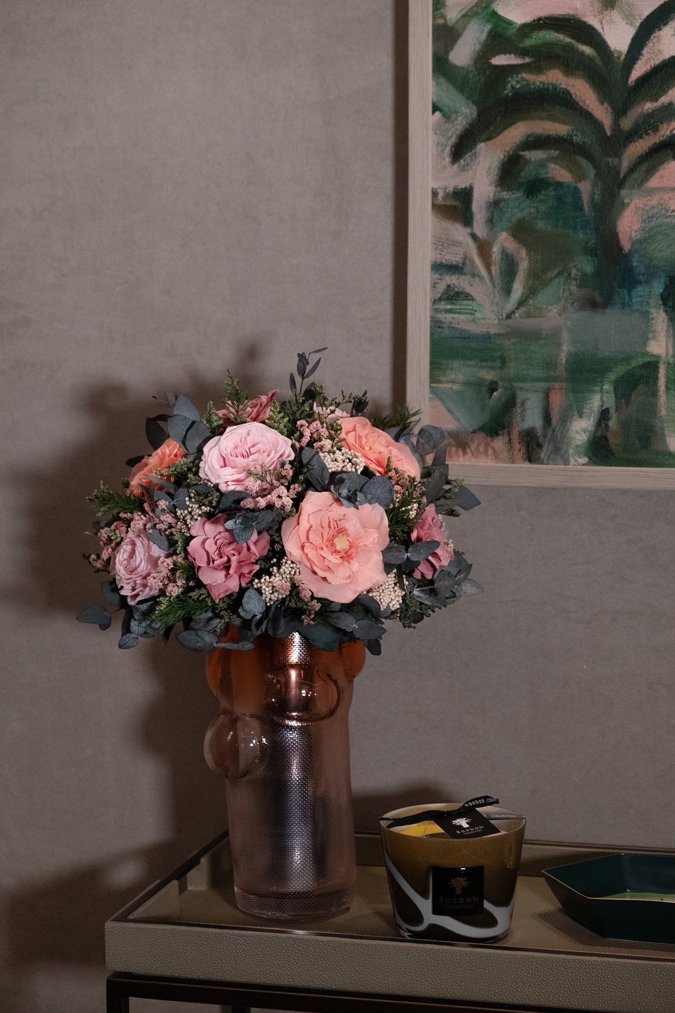 Bouquet of preserved pink flowers in a copper vase on a table with a painting in the background.