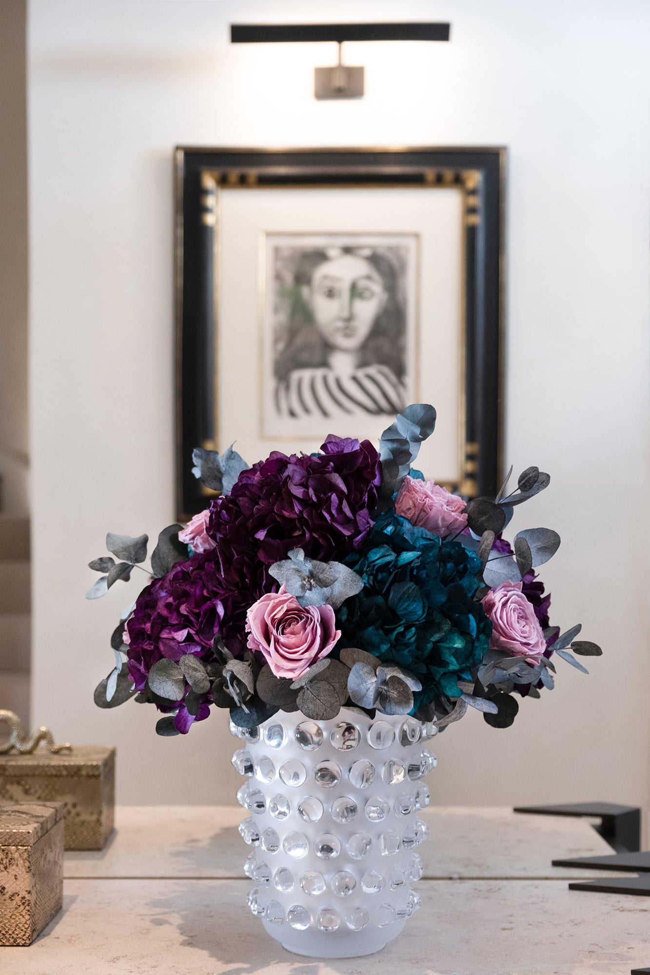 Decorative vase with preserved flowers on a table in a room with framed artwork.