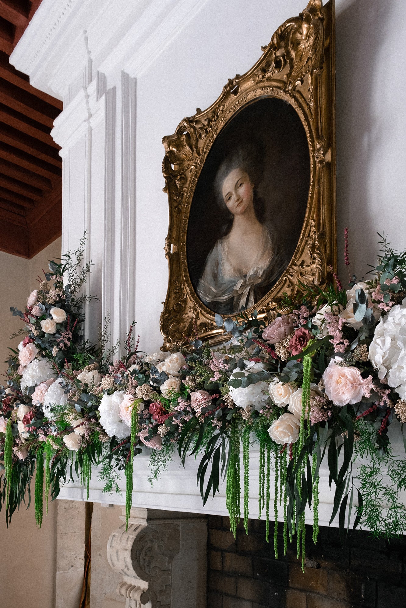 Preserved floral arrangement on a mantelpiece with a classical portrait in a gold frame above.