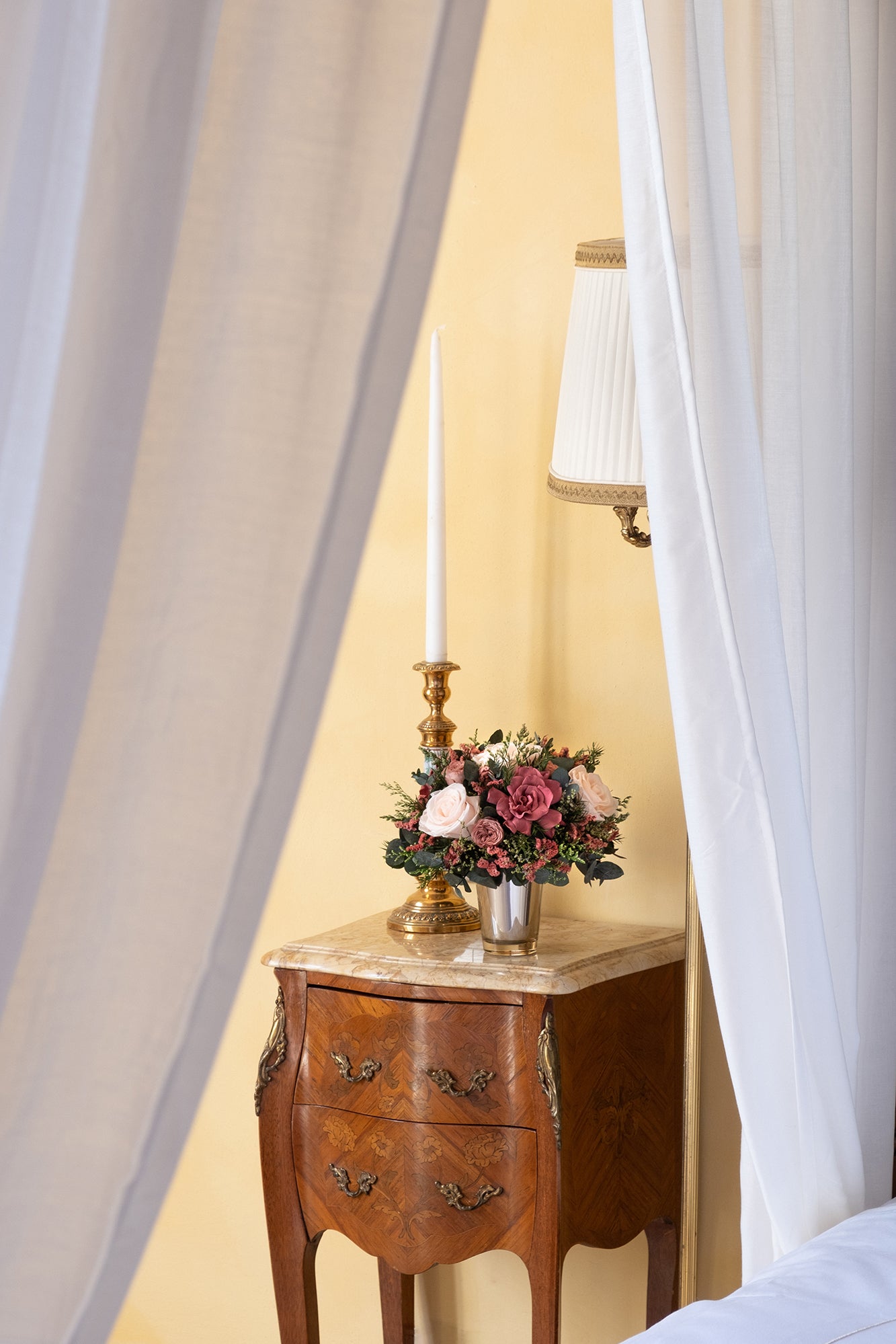 Wooden side table with a lamp and preserved mixed flower arrangement, draped with white curtains.