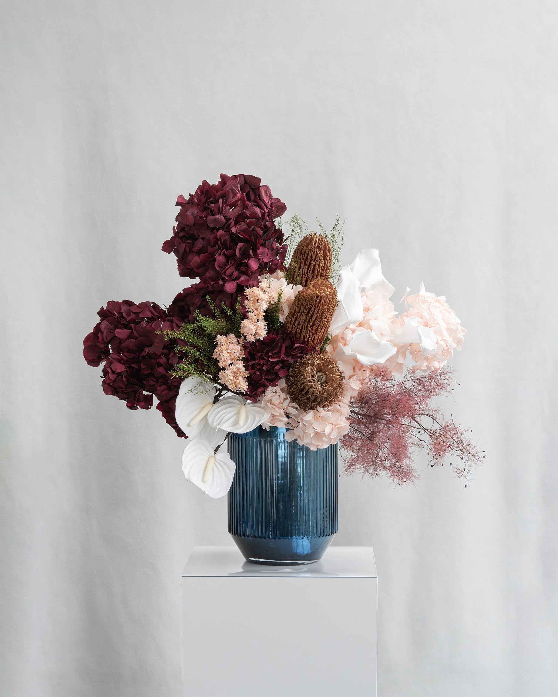 Preserved hydrangea, anthurium, starflower, and smoke bush arrangement in a blue vase