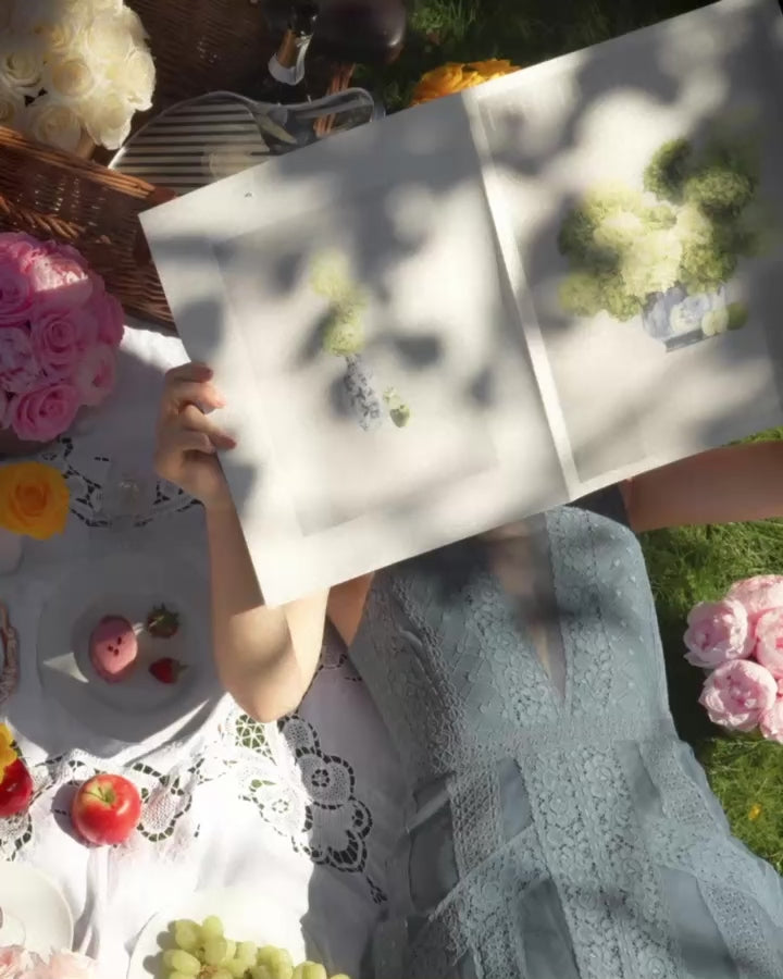 Protagonist surrounded by preserved flower arrangements reading a newspaper  whilst on picnic with a friend